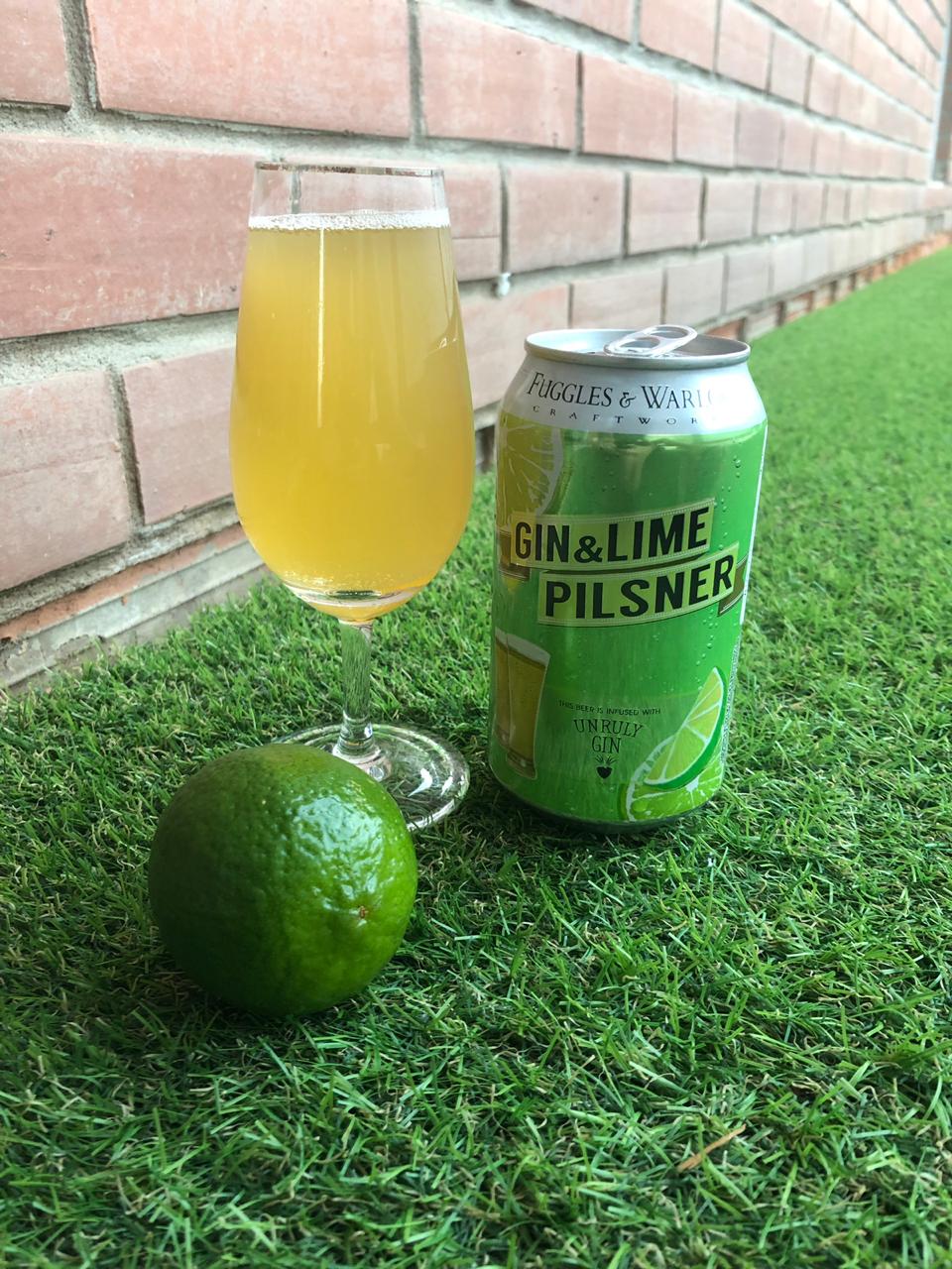 parede de tijolinhos chão com grama um limão ao lado de uma lata verde com cerveja de gin e limão e copo com cerveja clara