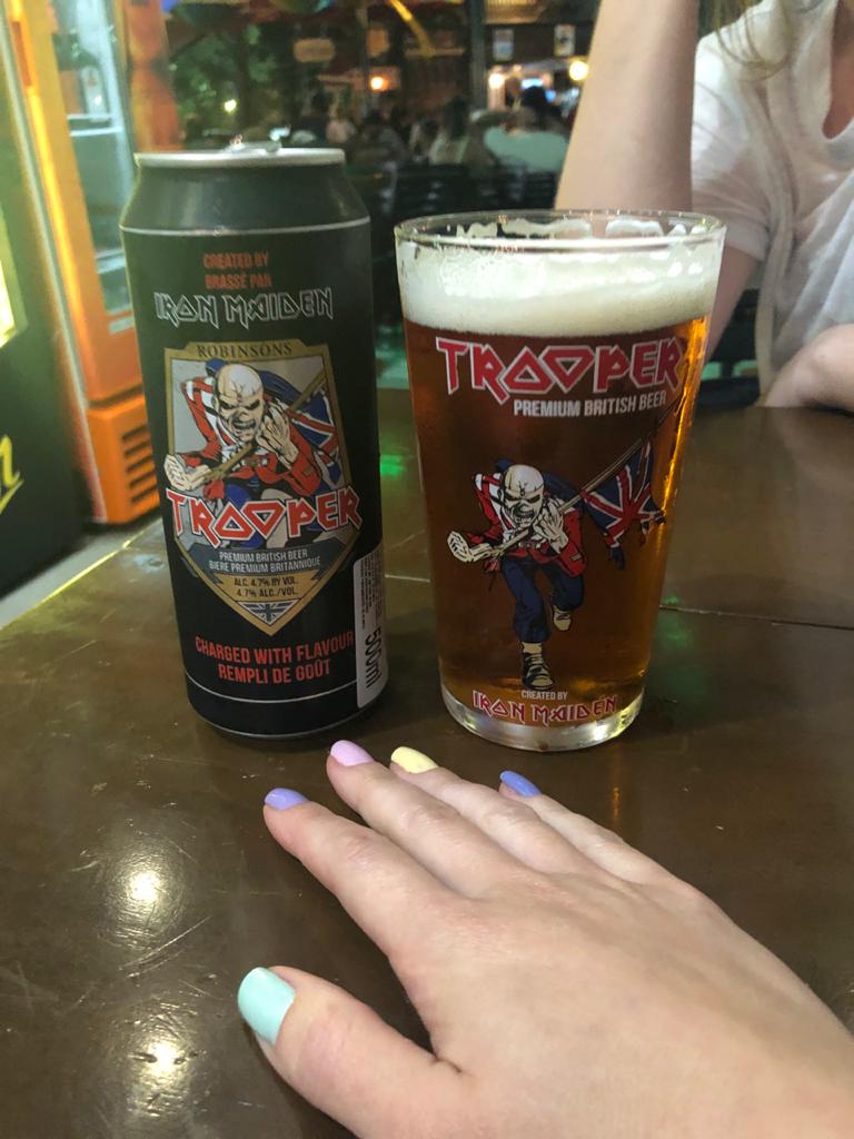 lata de cerveja e copo com cerveja Golden Ale do Iron Maiden em cima da mesa com mão pintada de esmaltes coloridos