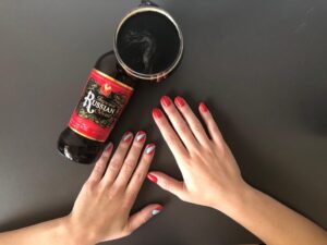 Garrafa de cerveja com rótulo vermelho Russian Imperial Stout copo com cerveja escura e mãos com unhas pintadas de vermelho e nail art vermelha e azul